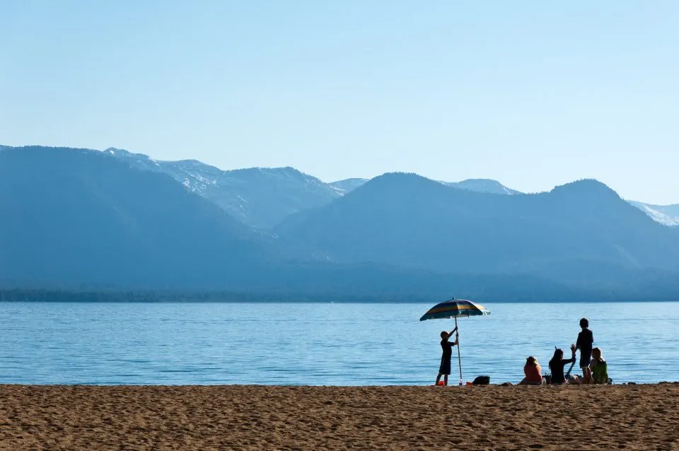 Família na beira de um lago com um guarda-sol, montanhas ao fundo